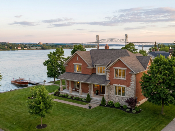 Aerial view of an elegant red brick waterfront home in Sault Ste. Marie featuring a lush lawn, private boat dock, and scenic river bridge view during golden hour.